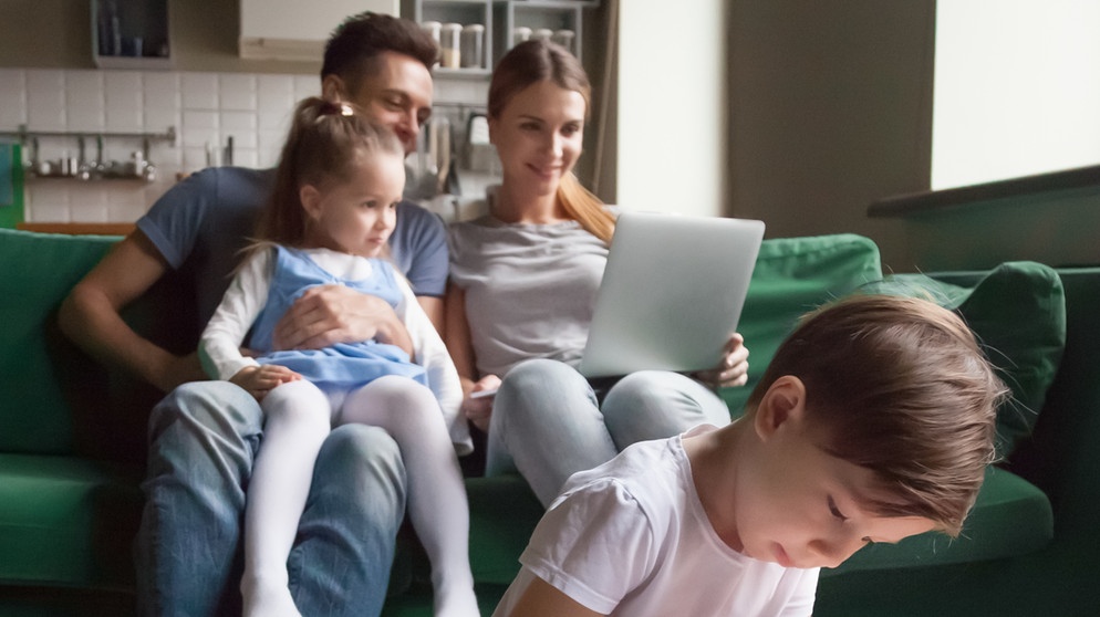 Vater und Mutter auf Couch zu Hause, Vater hat kleine Tochter auf dem Schoß, sie schauen in einen Laptop. Im Vordergrund mal der Sohn am Couchtisch ein Bild. | Bild: stock.adobe.com/fizkes